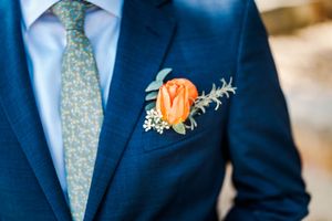Close-up of orange rose boutonniere with greenery on groom's navy blue wedding suit lapel.