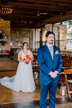 Bride in white gown holds orange bouquet as groom in blue suit stands for first look at rustic wedding venue.