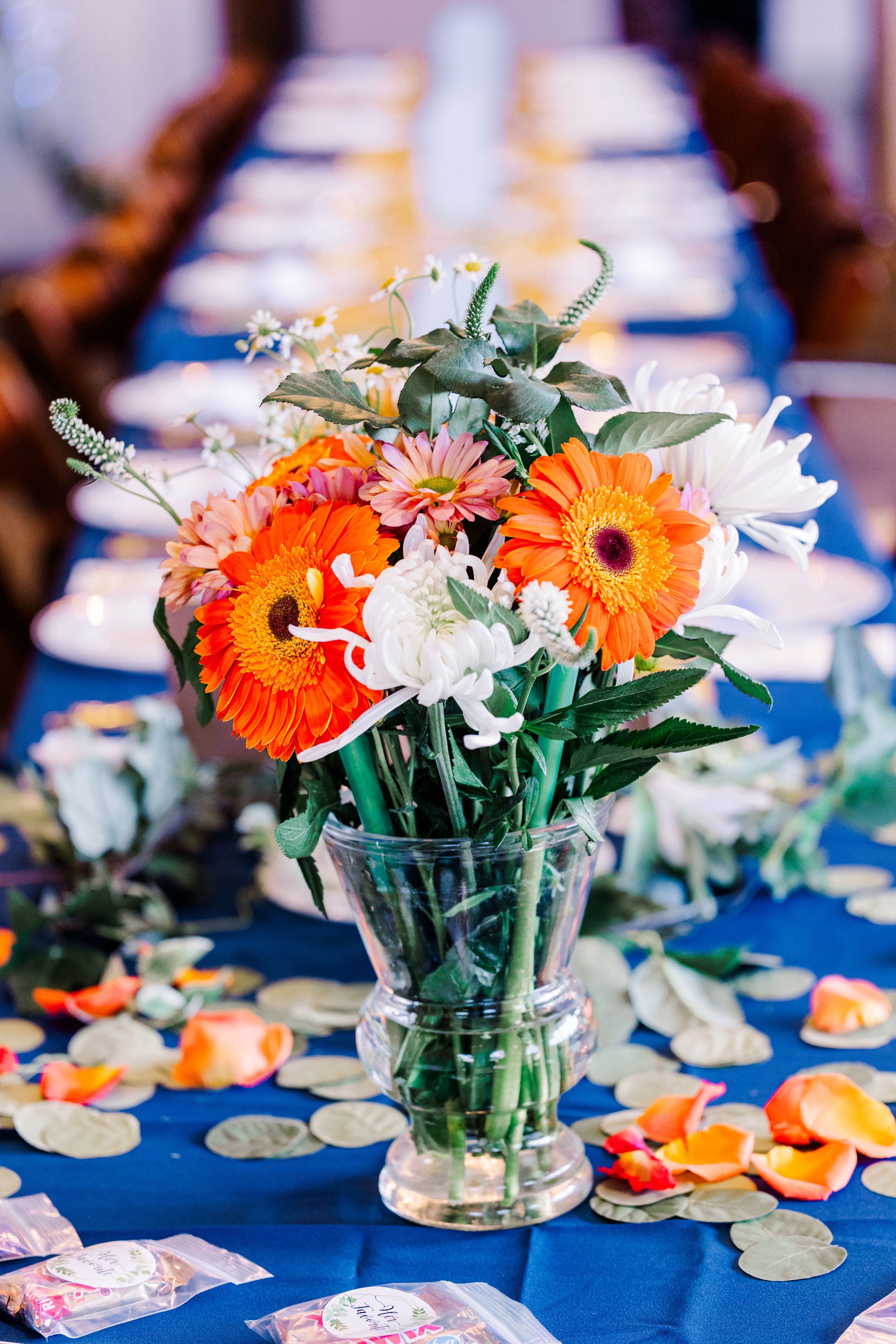 Orange gerbera daisy and white chrysanthemum centerpiece in glass vase on blue tablecloth.