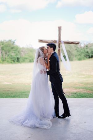 Bride and groom share first kiss at outdoor wedding ceremony with rustic wooden cross backdrop.