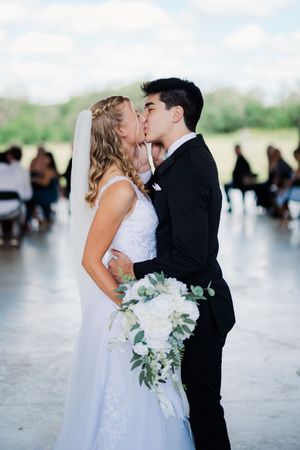 Newlyweds share romantic first kiss at wedding ceremony, bride holding white floral bouquet.