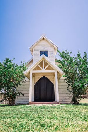 Charming small white wooden chapel with arched door surrounded by green trees and blue sky.