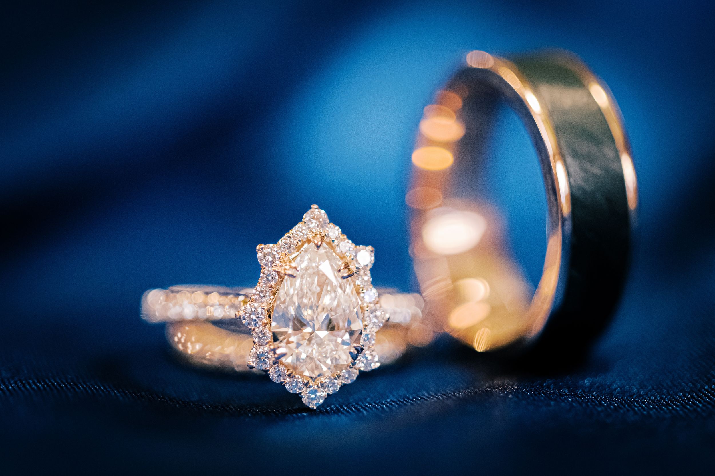 Close-up of marquise diamond halo engagement ring and gold wedding band on blue fabric.