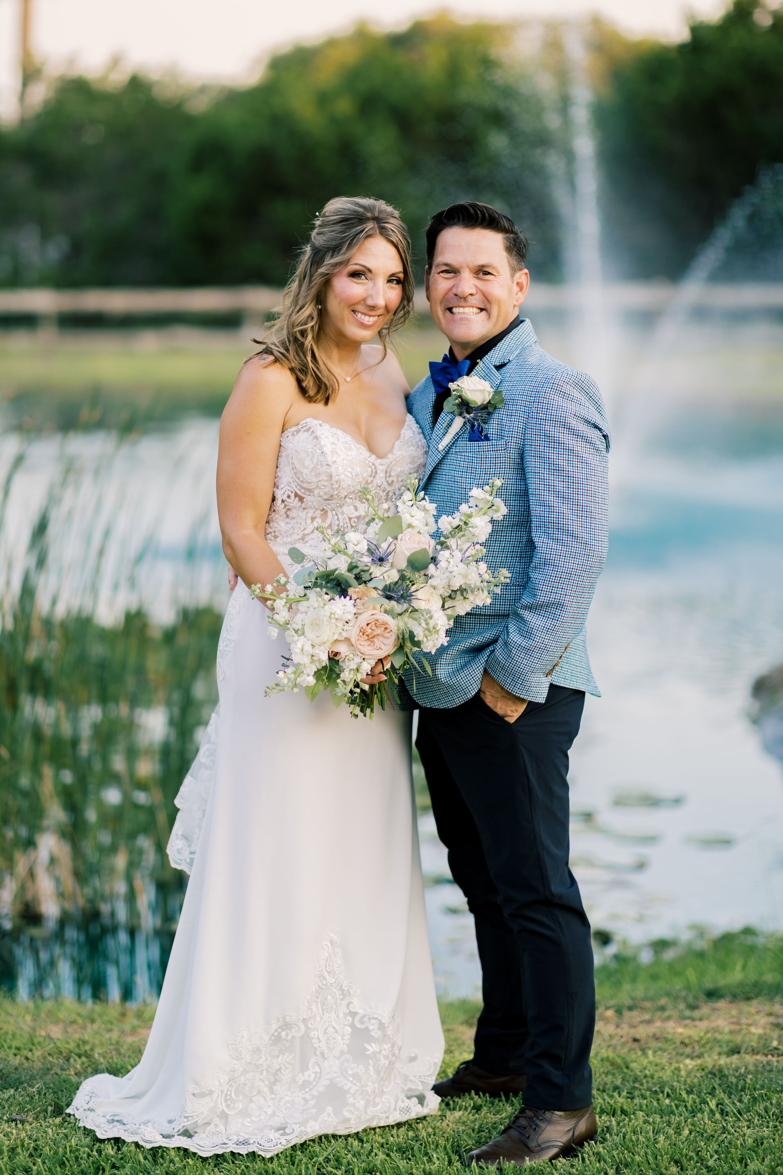 Smiling bride and groom pose together near pond with fountain at outdoor wedding venue.