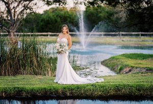 Elegant bride in fitted white gown poses alone near scenic pond with fountain backdrop.