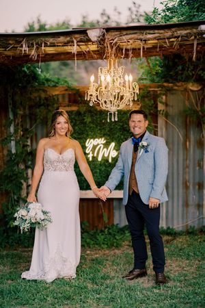 Bride and groom hold hands under chandelier with glowing neon you and me wedding sign.