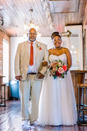 Bride in tulle ball gown walks with escort holding pink floral bouquet indoors.