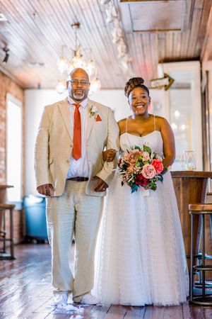 Bride in white tulle gown with pink bouquet walks with escort in tan suit and red tie indoors.