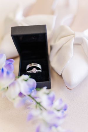 Diamond engagement ring in black box beside white bow heels and purple flowers.