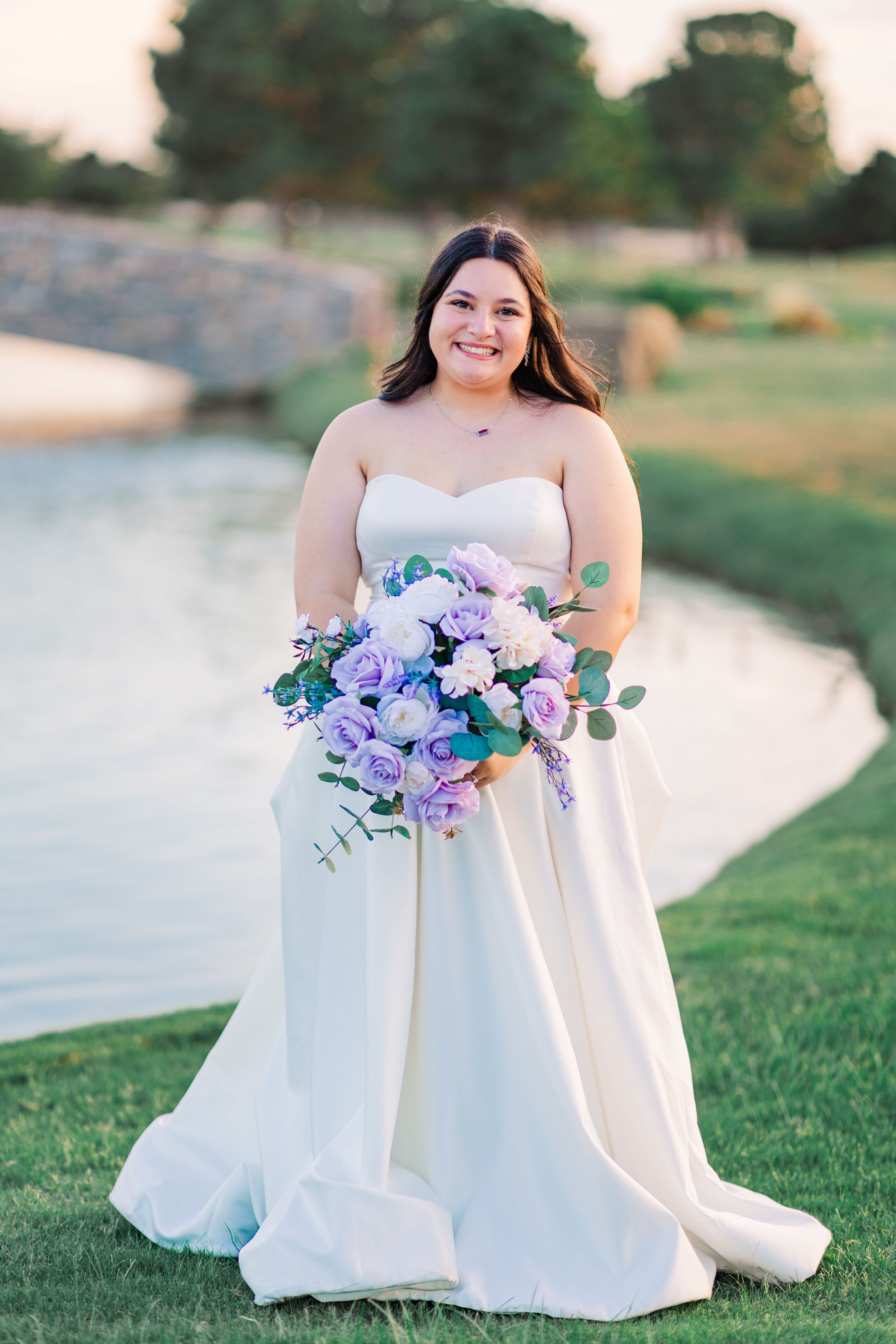 Smiling bride in strapless white gown holding purple and lavender bouquet outdoors near a pond.