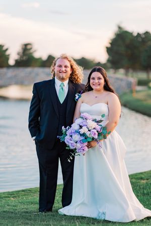 Bride and groom posing outdoors by a pond, she holds a purple bouquet, he wears a black suit.