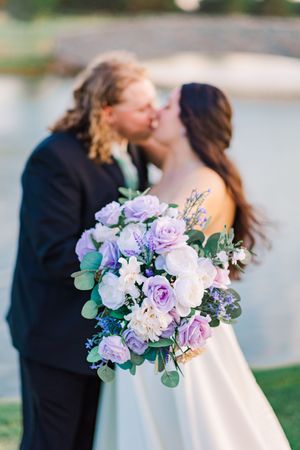 Couple kissing at outdoor wedding with blurred purple and white bouquet in foreground near pond.