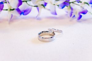 Two silver wedding rings displayed on white surface with purple and blue flowers in background.