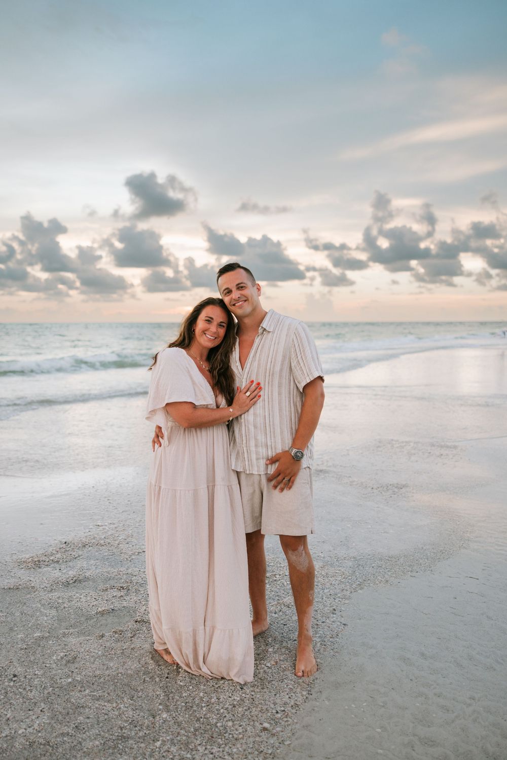 A couple shares tender moments together on a serene beach at sunset with soft cloudy skies in the background.