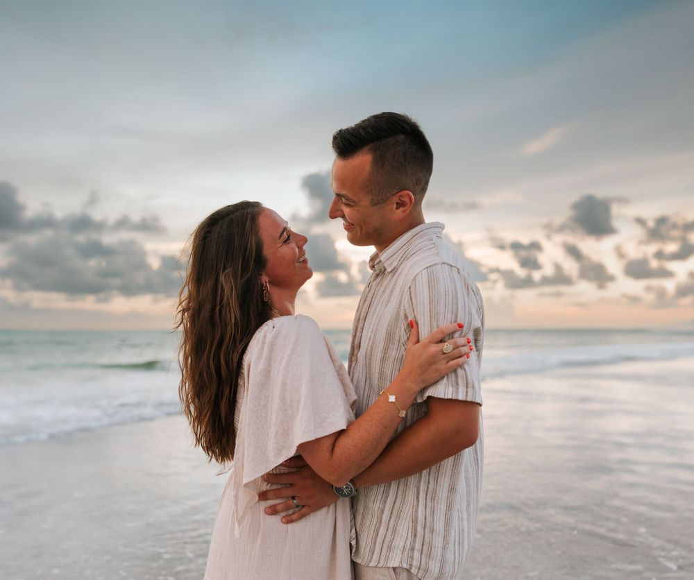 Couple embracing on sandy beach at sunset in matching white outfits creates romantic coastal scene.