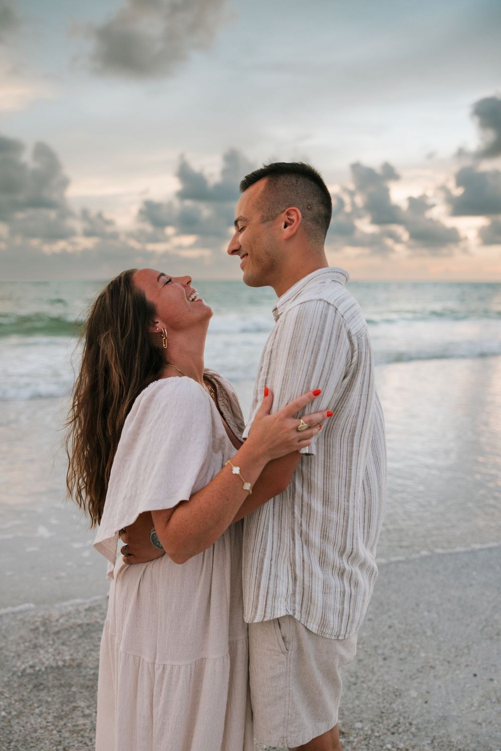 Romantic couple embracing on a beach at sunset with ocean waves and cloudy sky in the background.