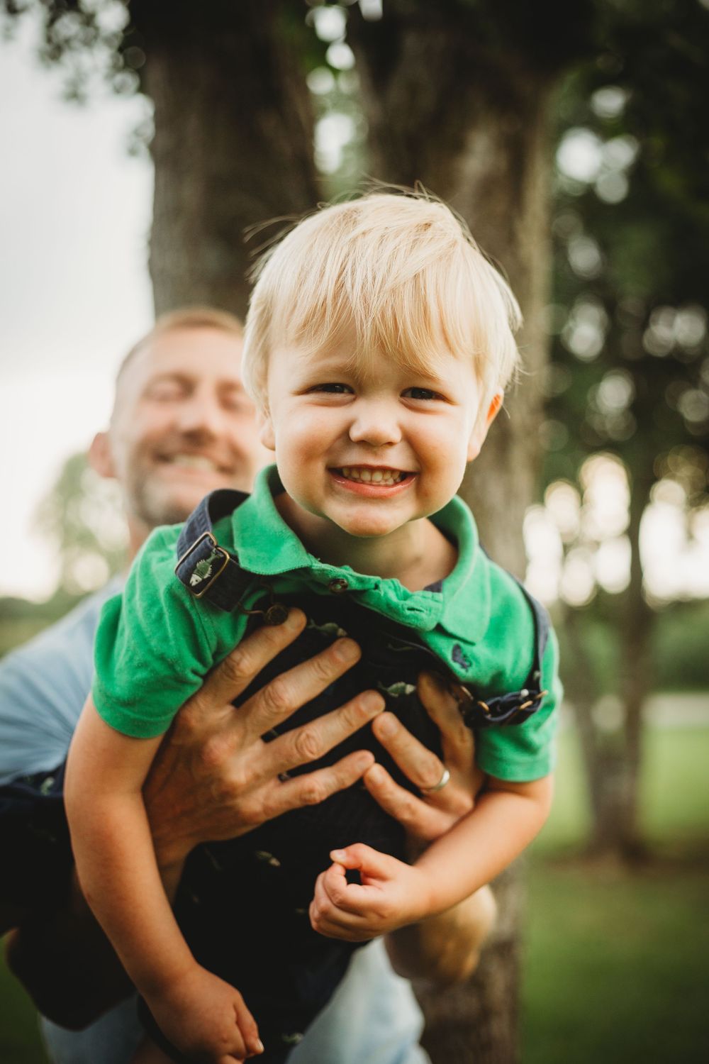 A child in a bright green shirt being playfully held up and swung around in a park setting.