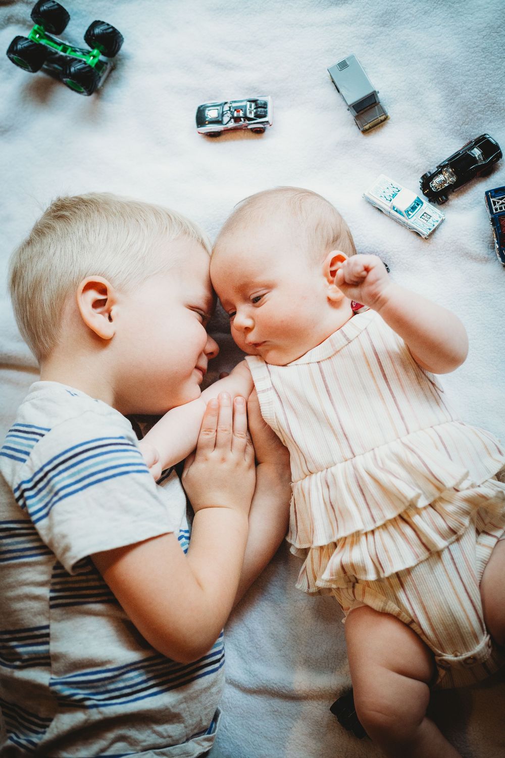 Two babies lying side by side, wearing light colored clothing, with small toy cars scattered nearby.