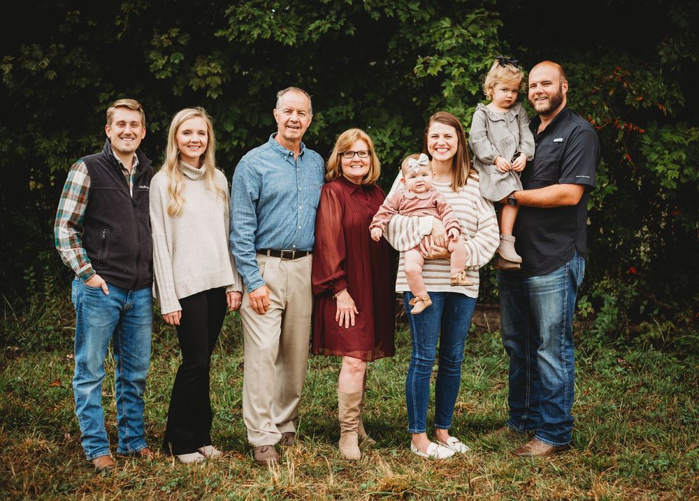 Multi-generational family standing together outdoors in casual attire against a natural foliage background.