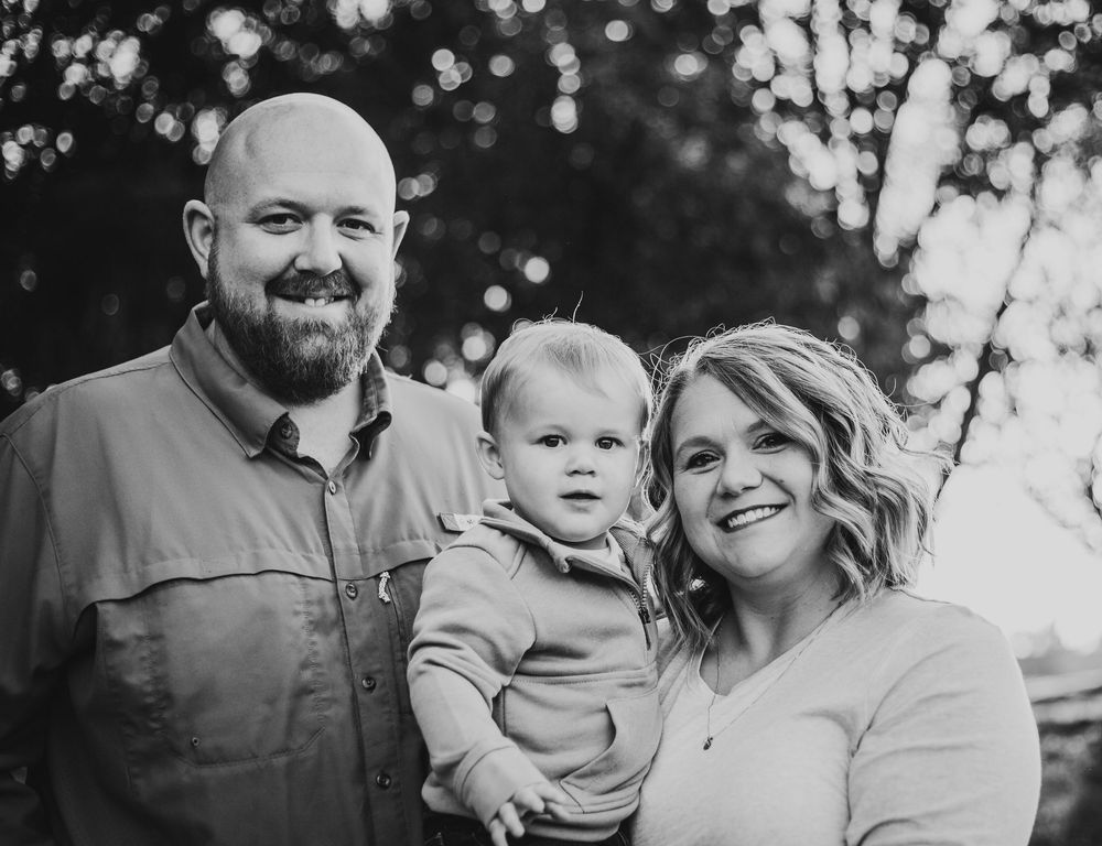 Black and white portrait of a family of three outdoors with bokeh lighting in the background.