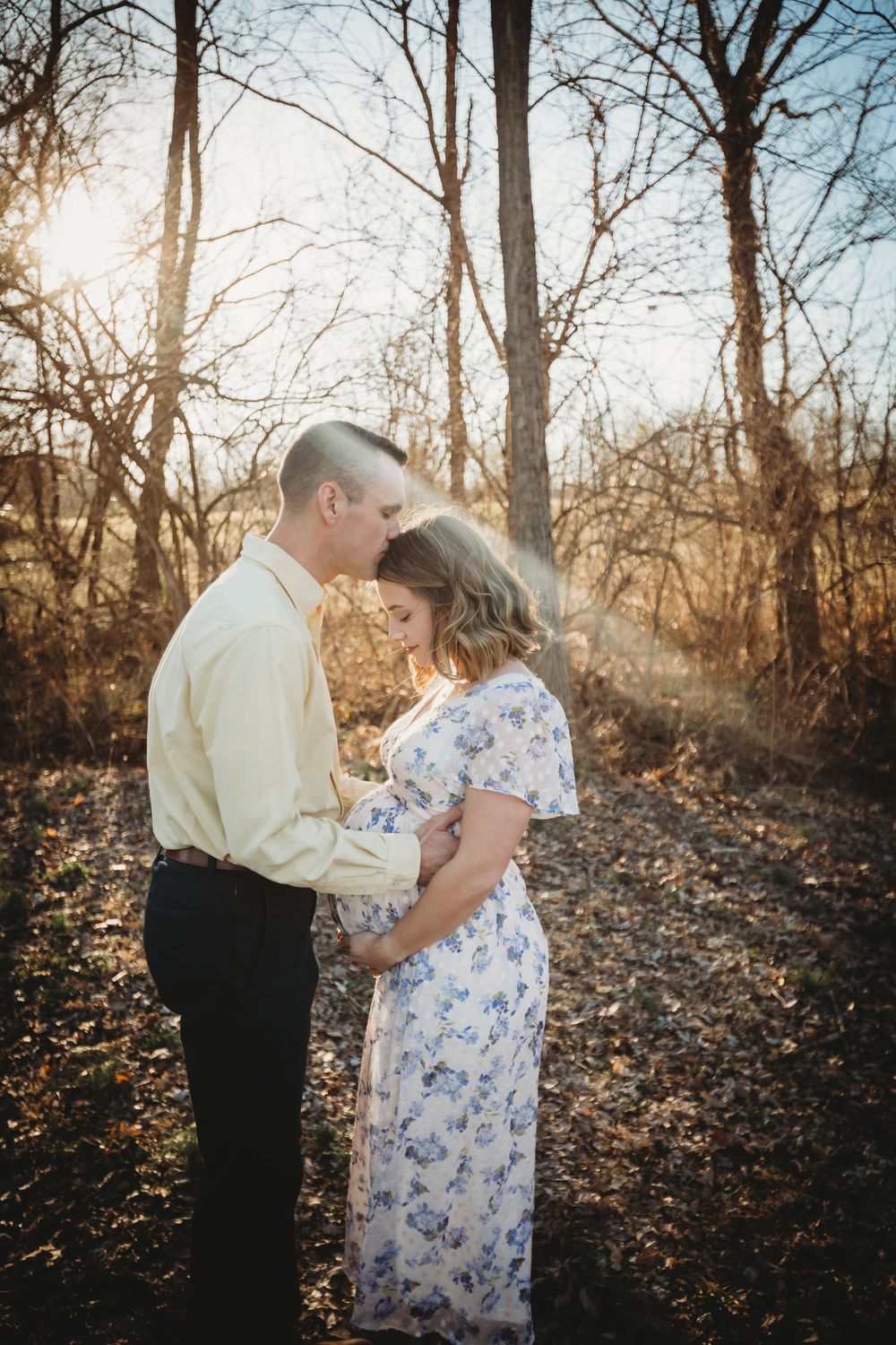 A romantic couple embraces in a sunlit forest during golden hour, with bare trees and fallen leaves around them.