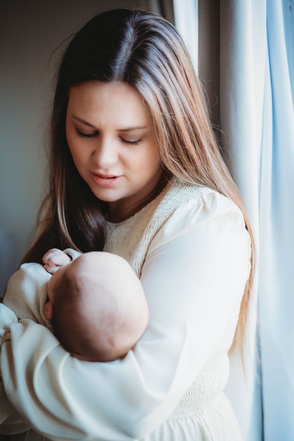 A tender moment between a mother in a white sweater holding her sleeping newborn baby near a window.