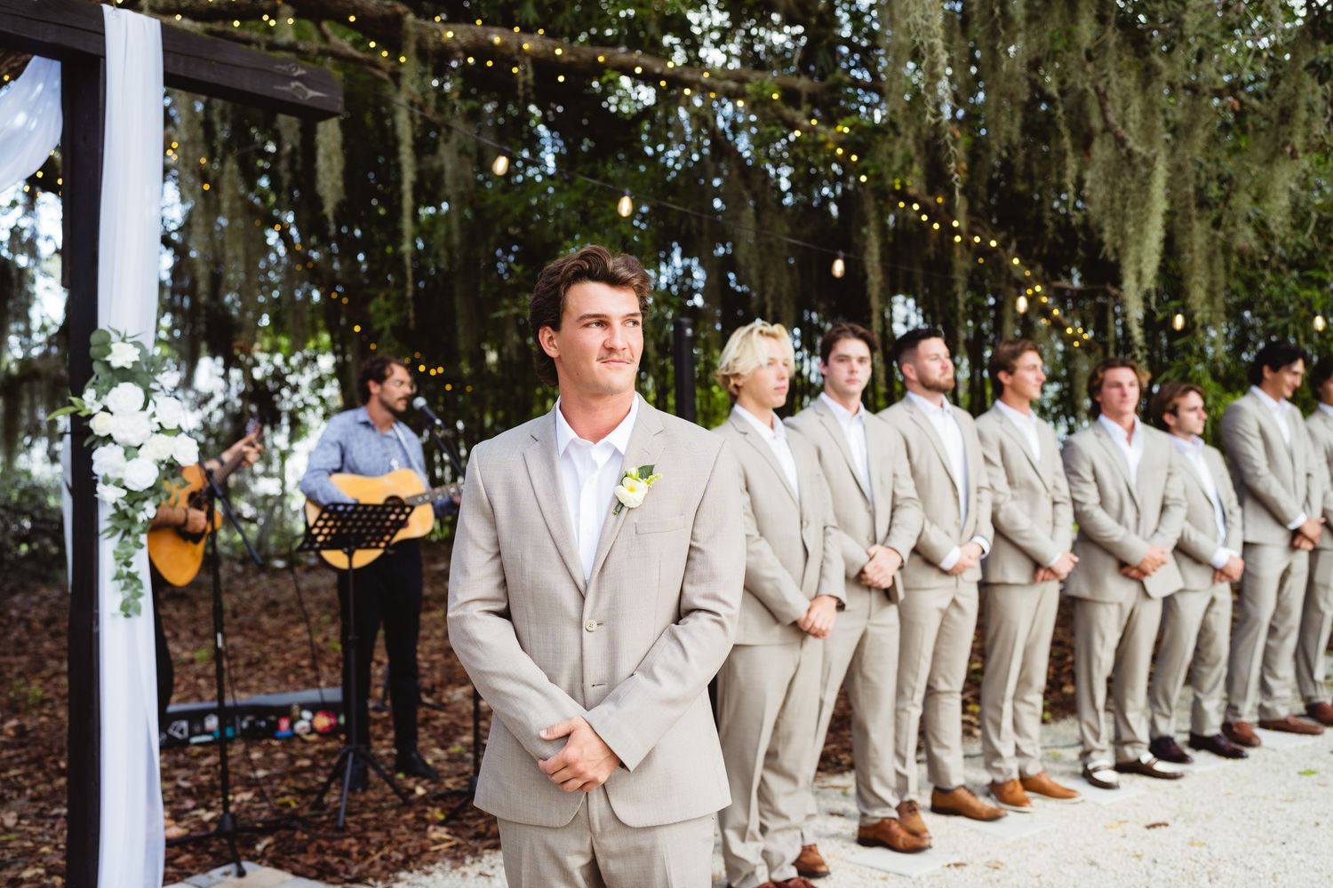 Wedding party line of groomsmen in light beige suits lined up outdoors with twinkling lights in trees.