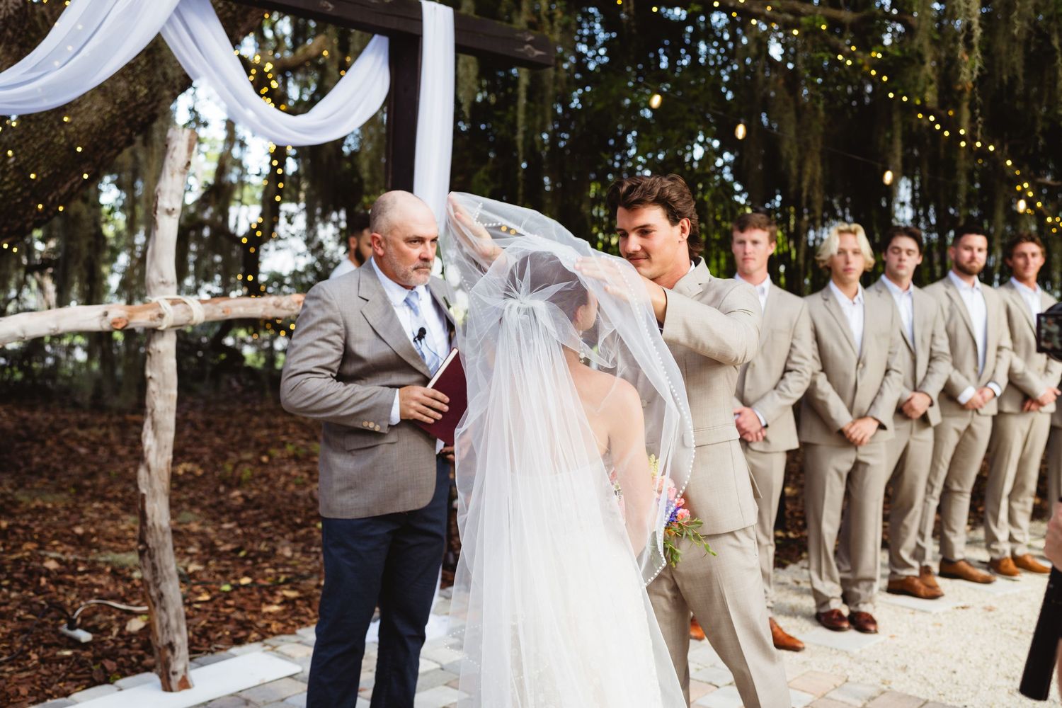 Groomsmen and bridesmaids pose in line formation at outdoor wedding under string lights and oak trees.