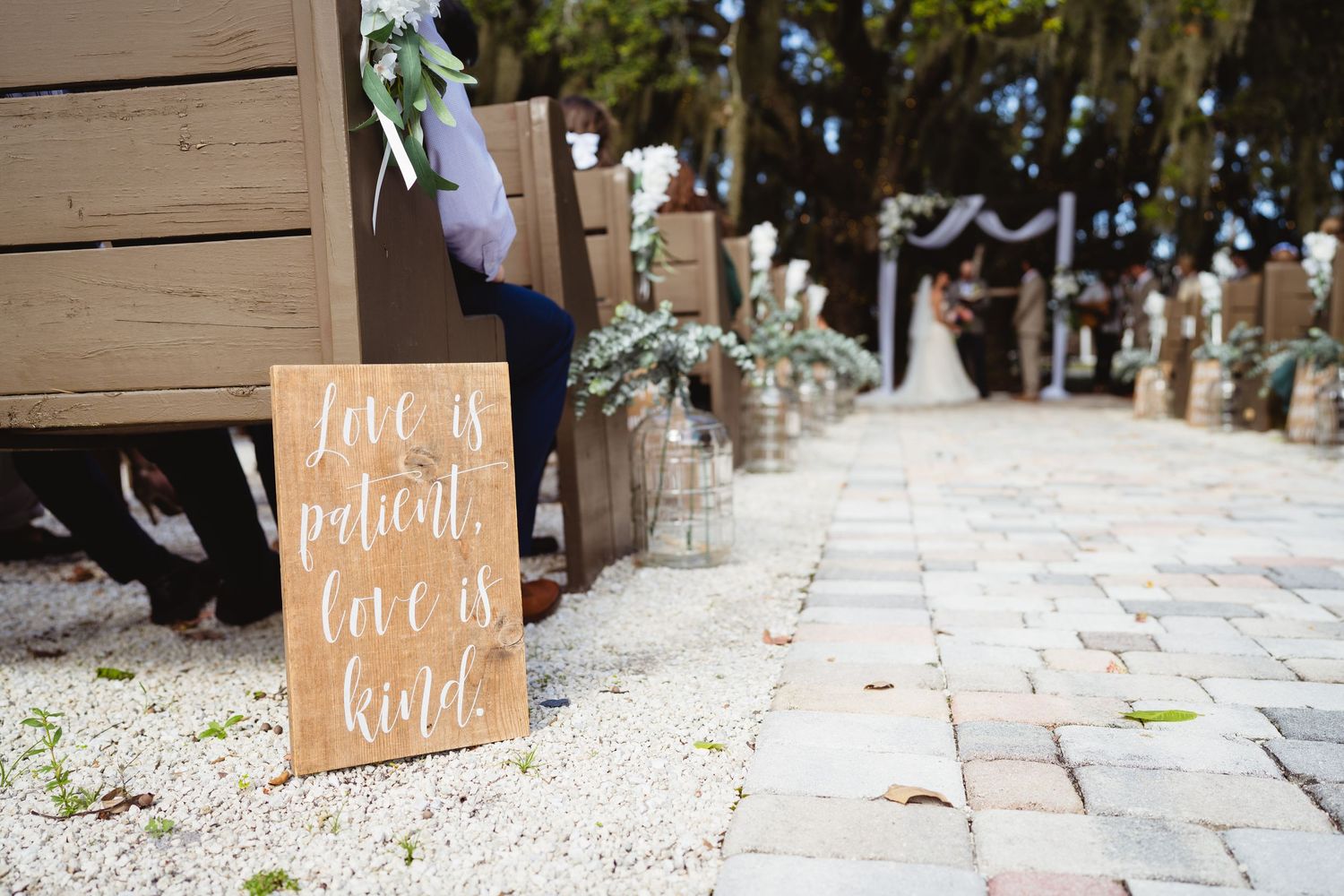A rustic wooden wedding sign with jars of flowers lines a stone pathway at an outdoor ceremony venue.