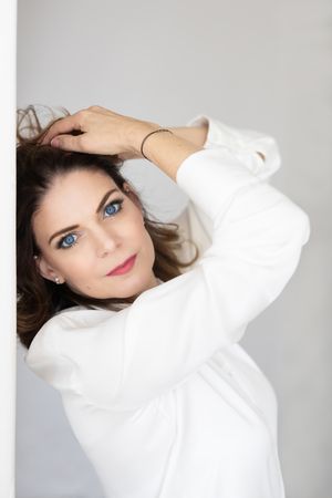 A woman with blue eyes wearing a white sweater poses against a light gray background while styling her dark hair.