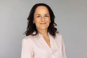 Person wearing soft pink blouse smiles warmly against a neutral gray background in professional headshot.
