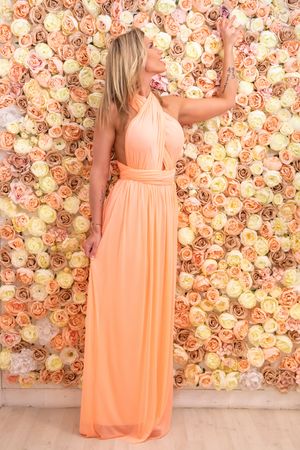 A peach-colored flowing maxi dress worn against a romantic floral wall backdrop of blush and cream roses.