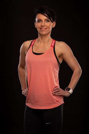Athletic person in coral tank top and black workout pants poses against dark background with smartwatch and fitness attire.