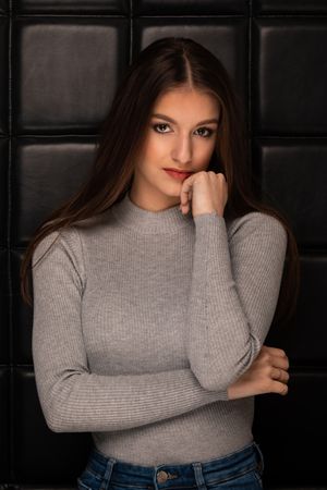 Woman in gray ribbed sweater and jeans poses thoughtfully against dark tiled wall in moody portrait lighting.
