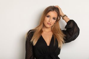 A woman poses against a white wall wearing a black wrap dress with sheer long sleeves.