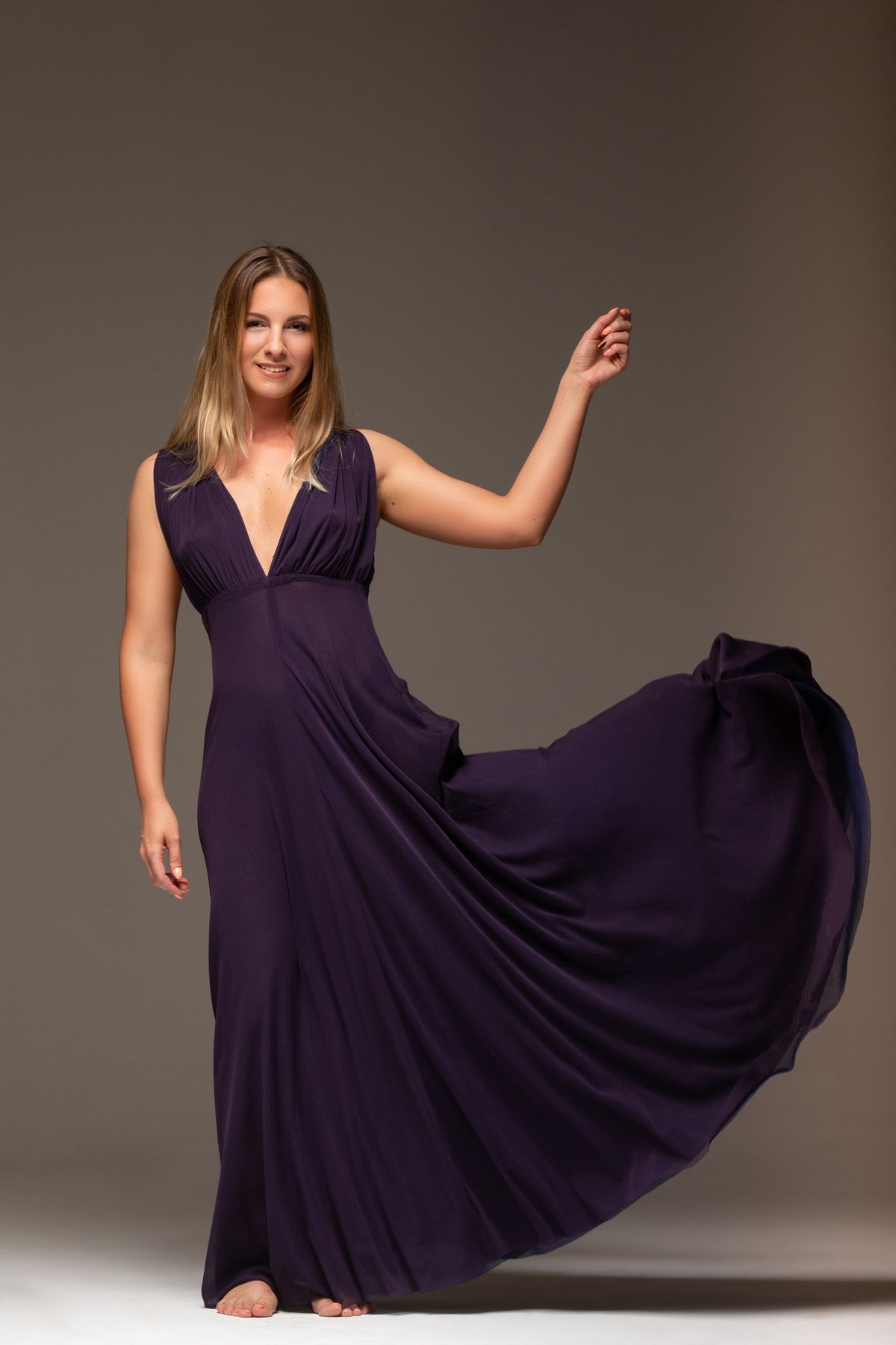 A flowing dark purple maxi dress with a plunging neckline and full skirt twirls elegantly against a gray studio background.