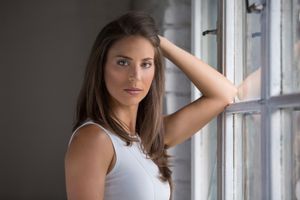 Woman in white tank top standing by window with natural light, hand in long brown hair.