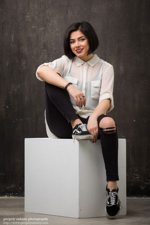 Portrait of someone in casual attire with white shirt and ripped black jeans sitting on a white cube against dark background.