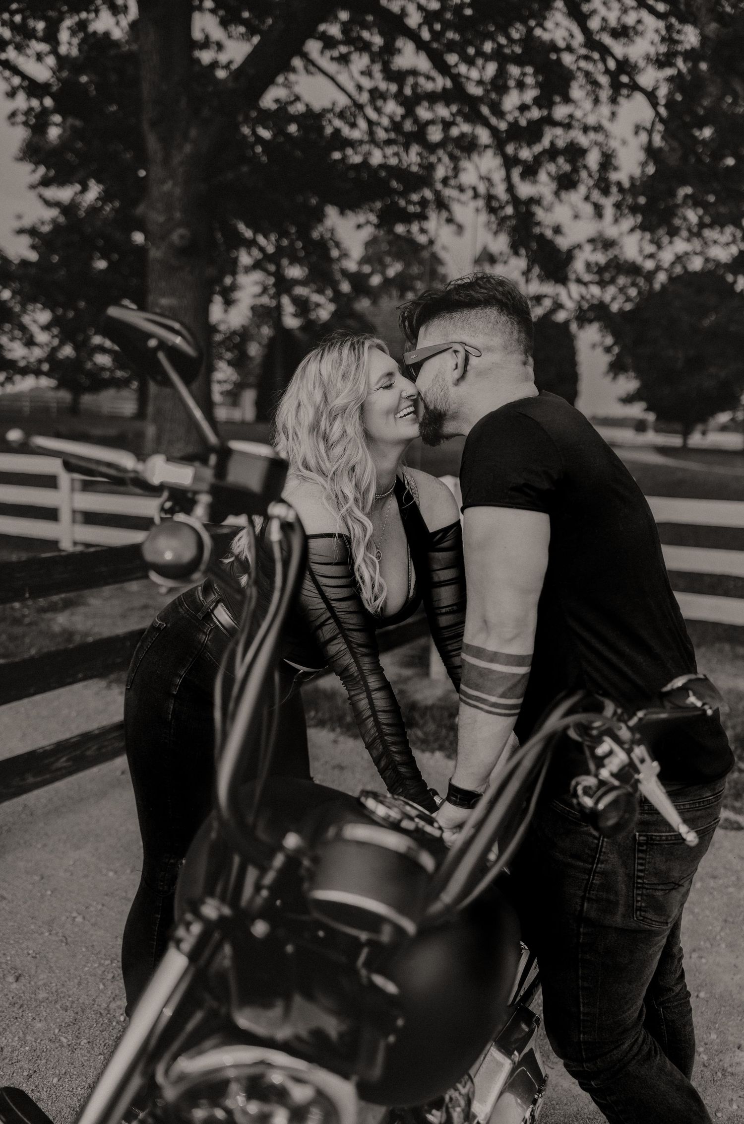 A romantic motorcycle photoshoot in black and white features a couple embracing beside a vintage bike on a tree-lined path.