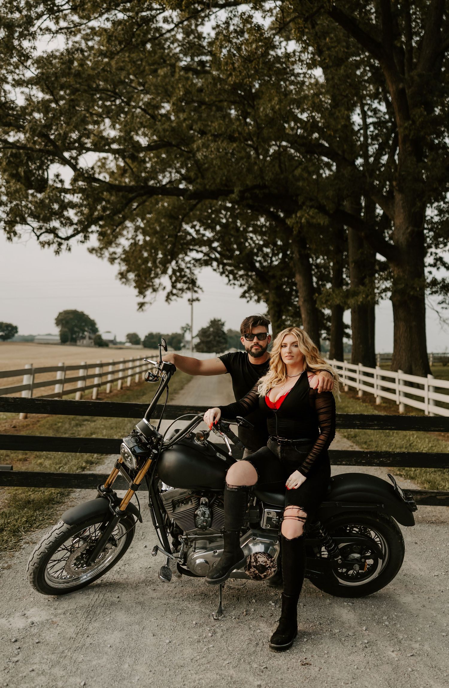 A couple poses with their classic motorcycle on a country road with white fence and trees in the background.