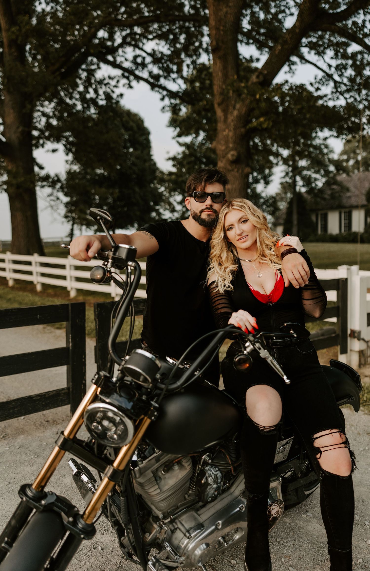 A couple in edgy black outfits poses with their motorcycle against a rustic farm backdrop.