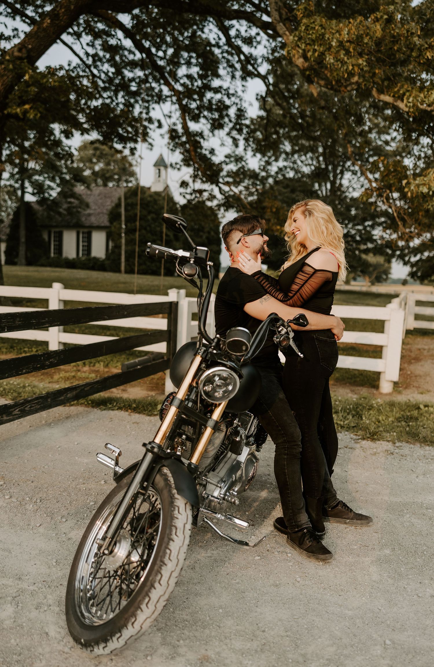 A couple shares a tender moment while leaning against their motorcycle on a country road.