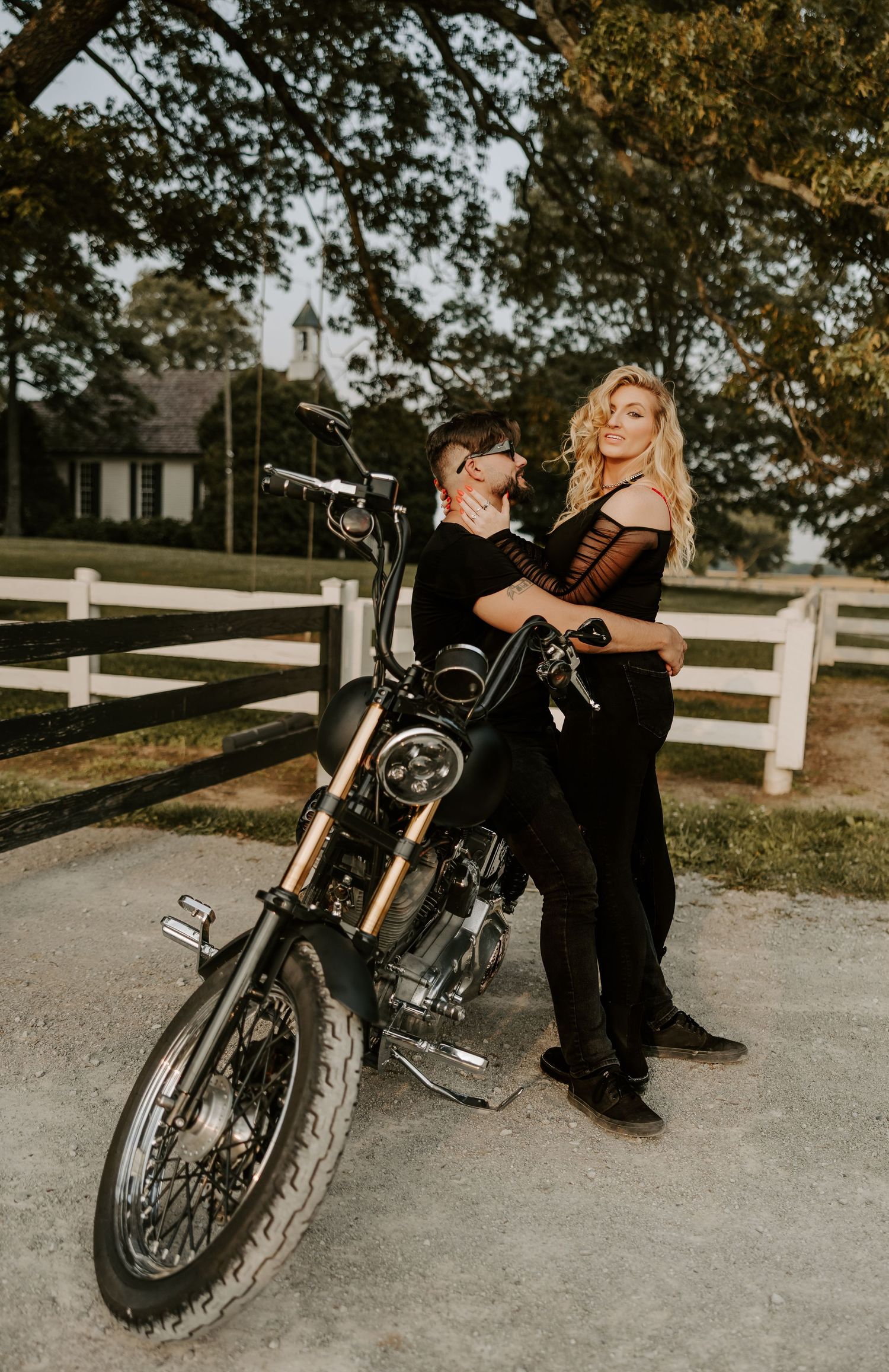 A romantic motorcycle engagement session features a couple posing with their bike near white fencing.