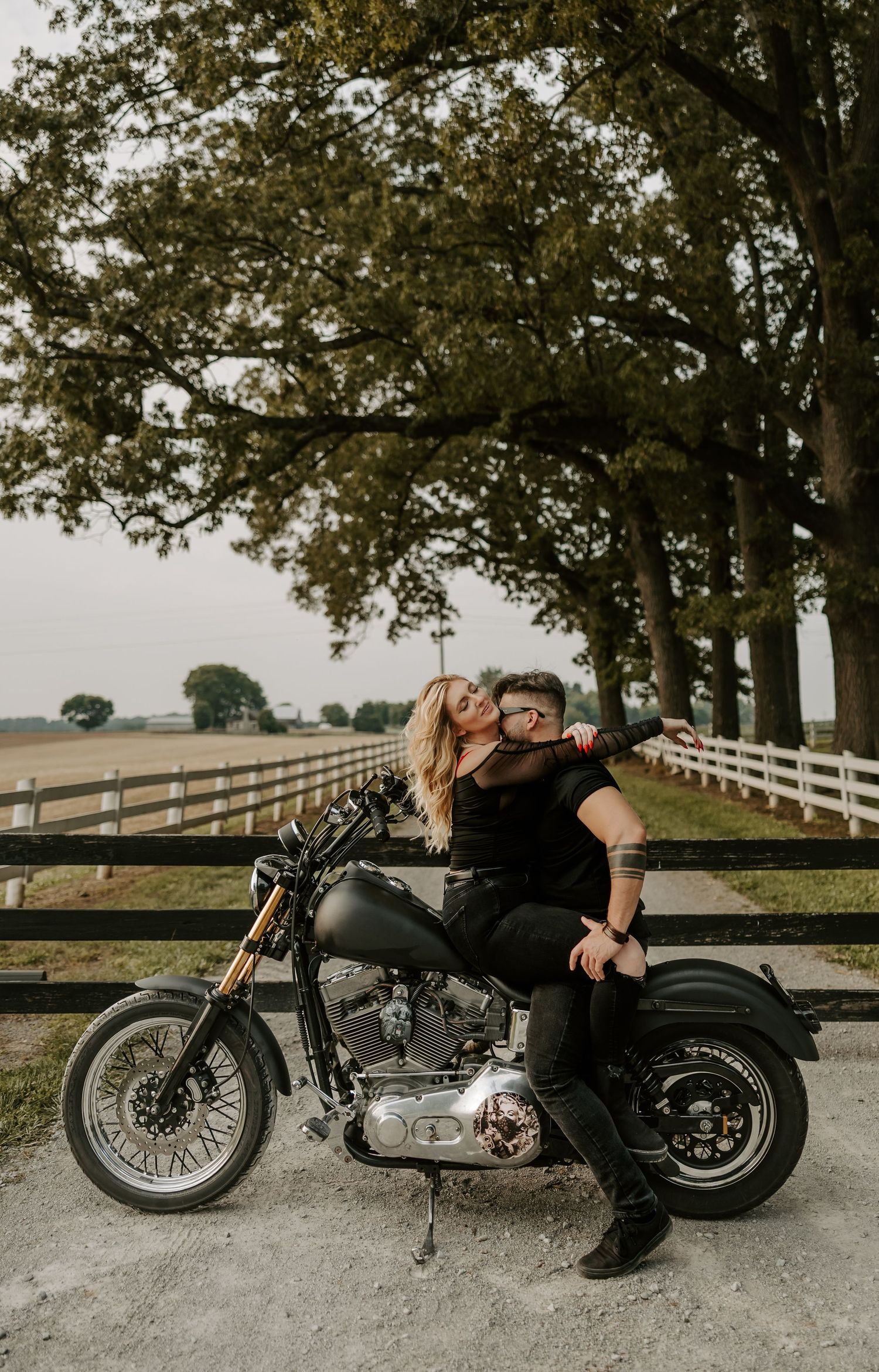 A romantic motorcycle photoshoot captures a couple embracing on their bike with a scenic farm backdrop.