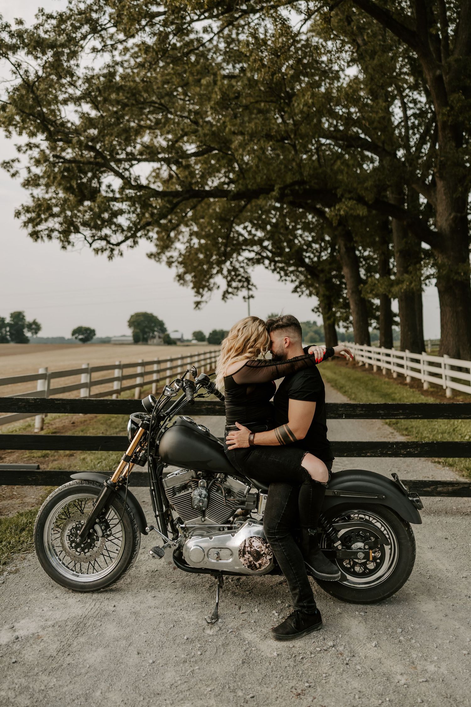A couple shares an intimate moment on their classic motorcycle against a backdrop of trees and white fencing.