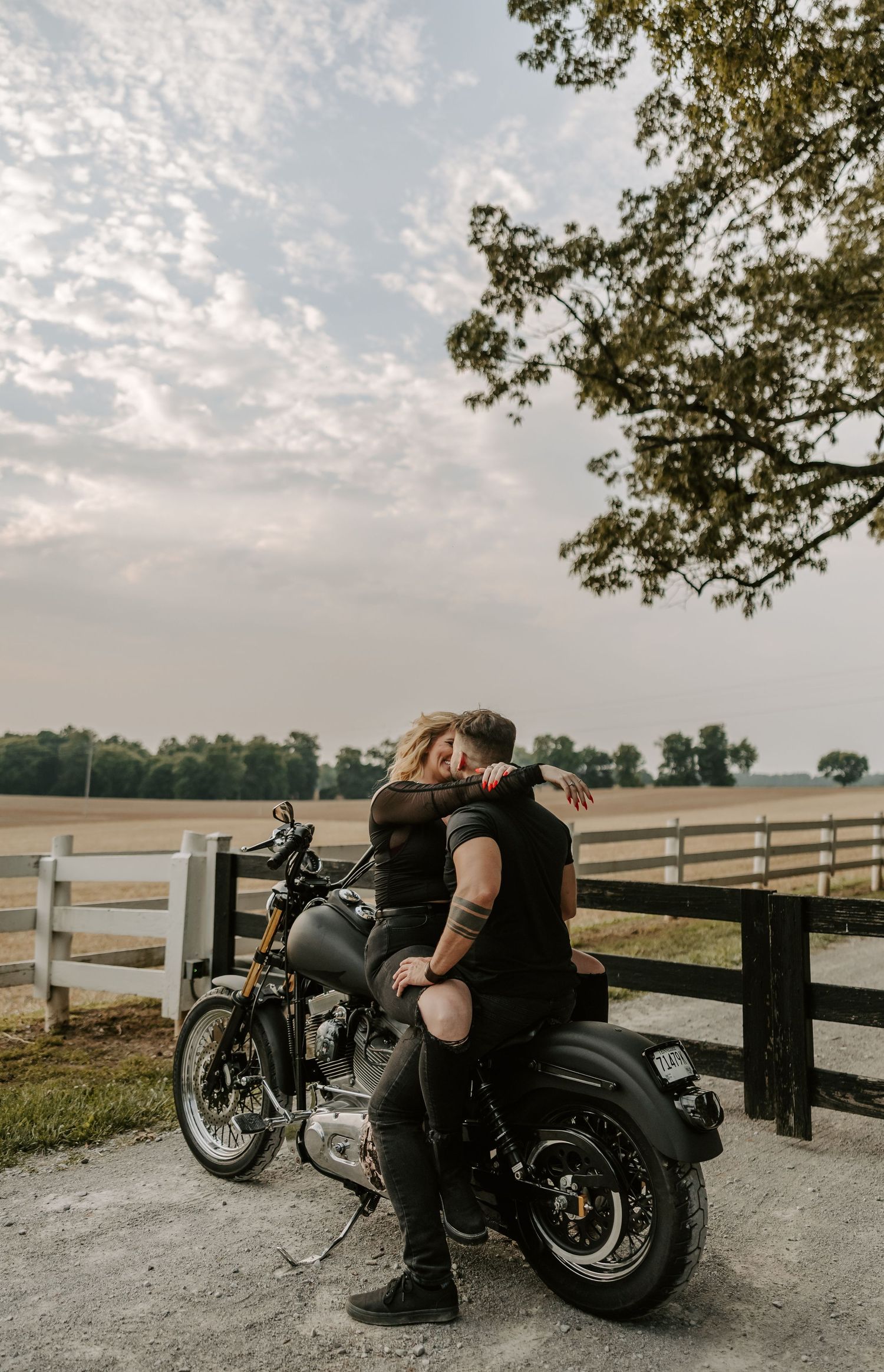 A sunset motorcycle engagement session shows a couple embracing by their bike with white fencing in background.