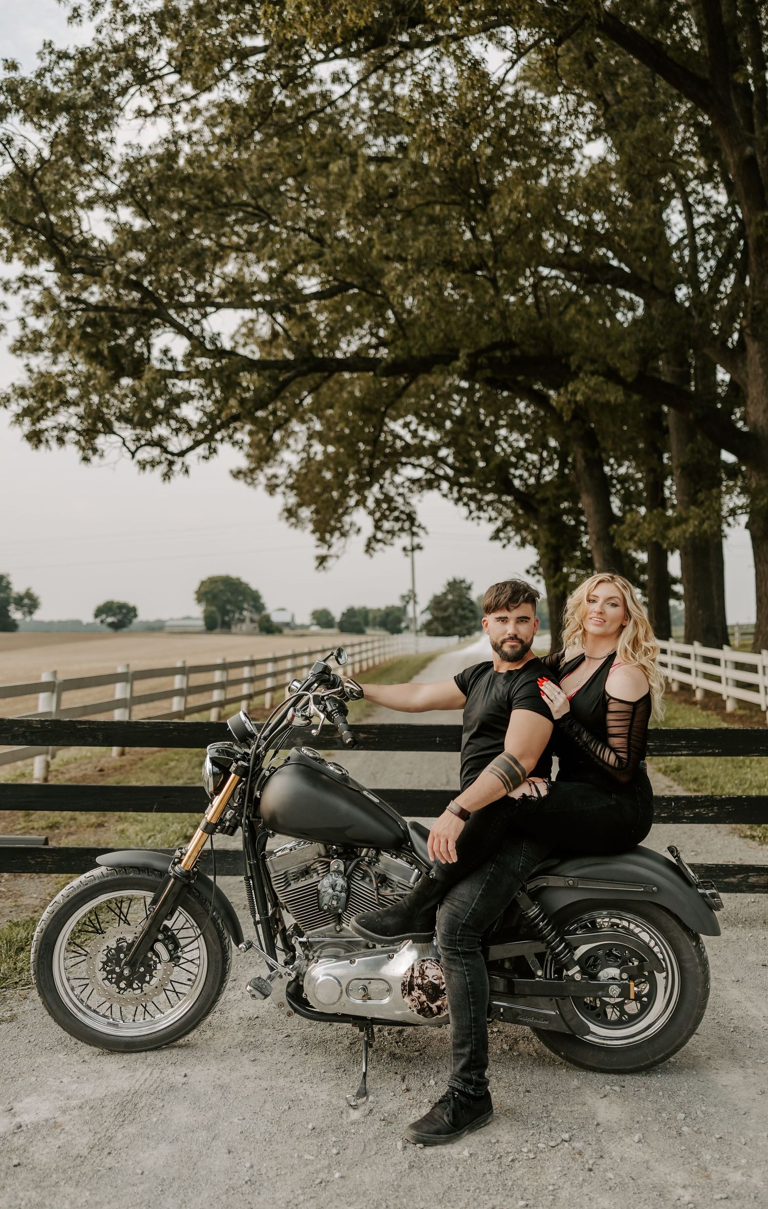 Black and white photo of couple lounging against classic motorcycle under trees.