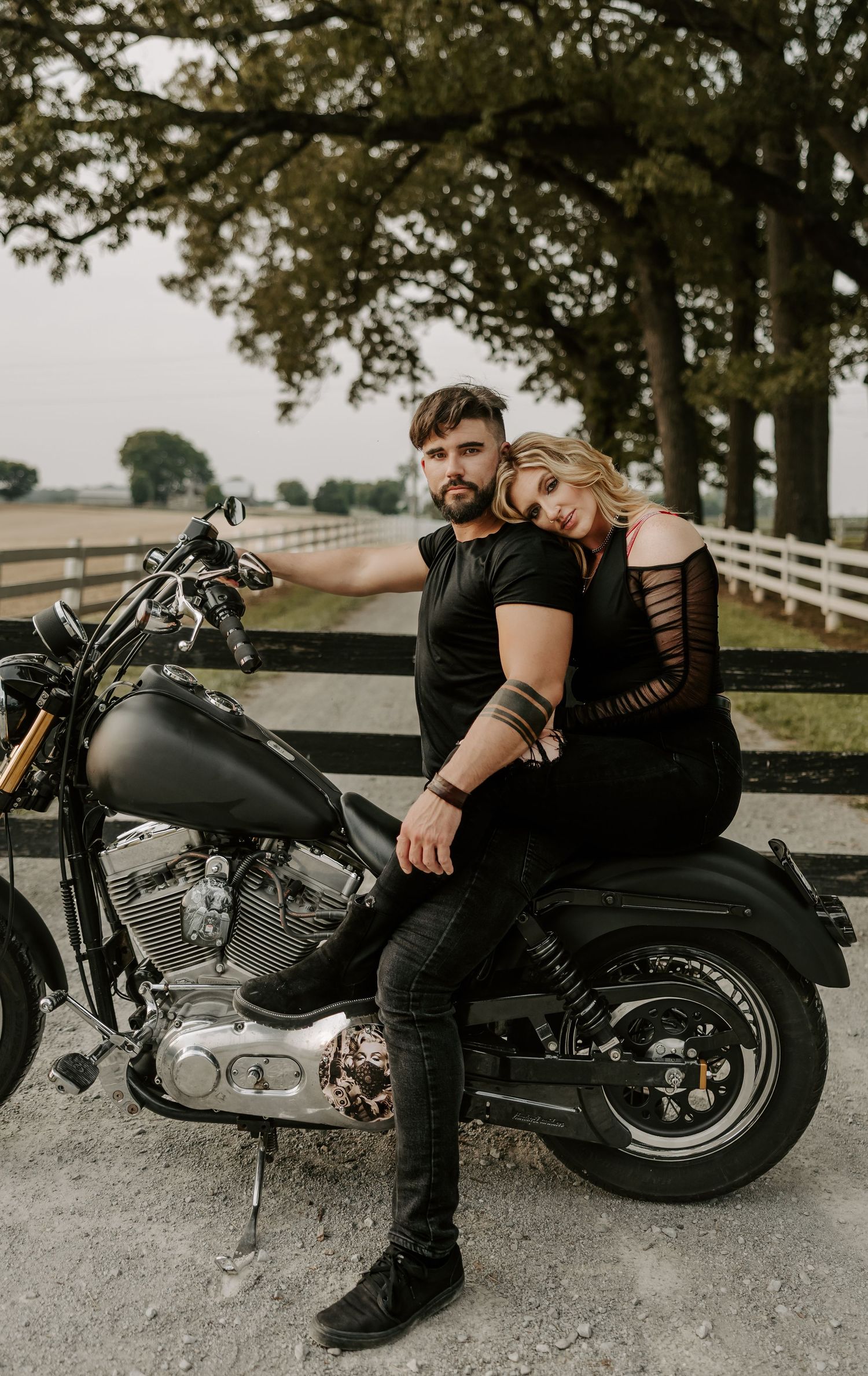 Couple in black clothing poses with custom motorcycle on gravel path near white fence.