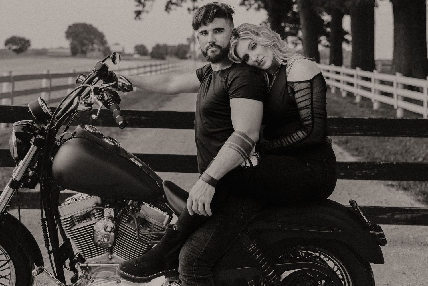 Romantic couple poses with vintage motorcycle on a country road with white fence and trees in background.