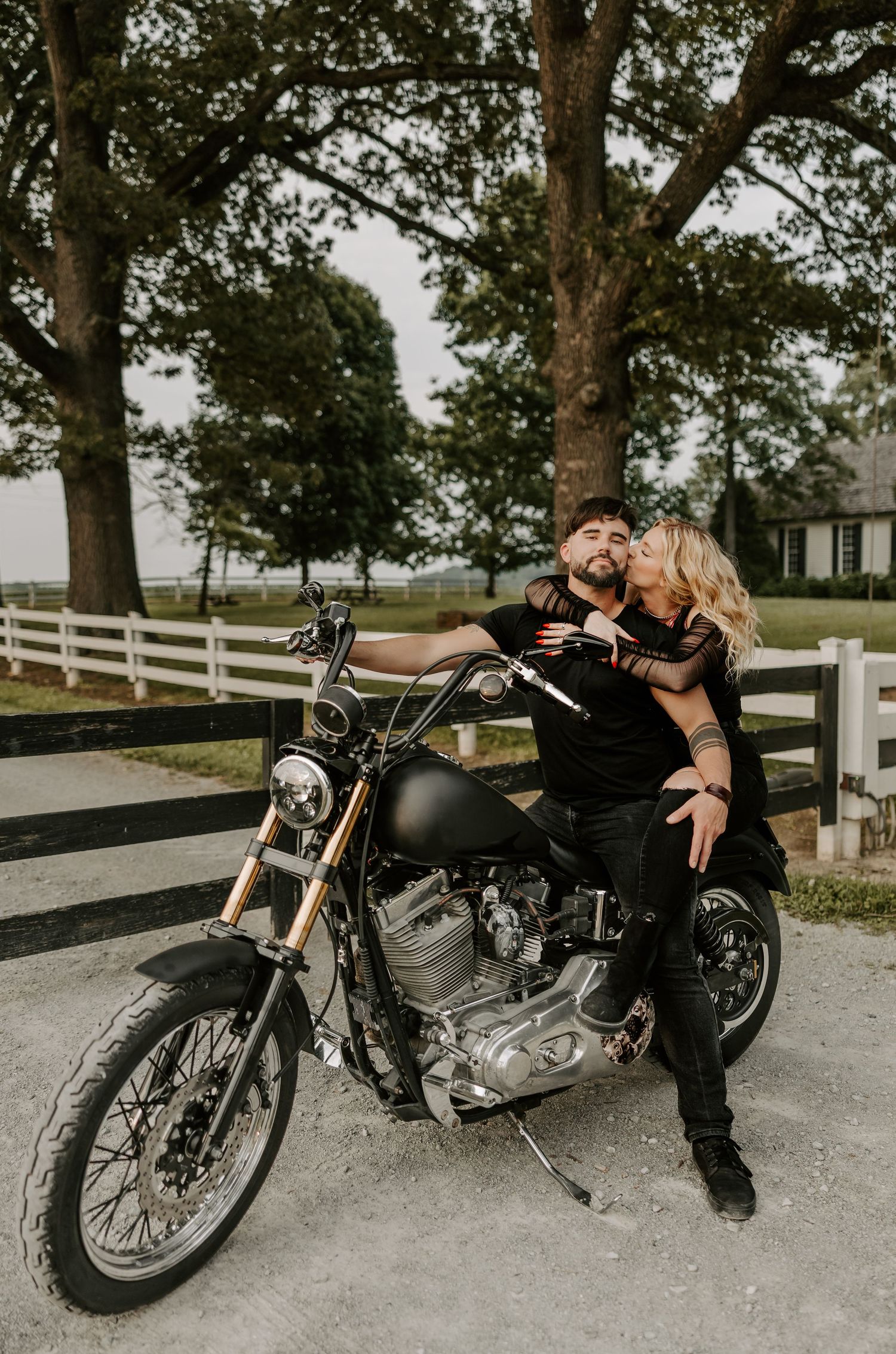 Couple lounges romantically on vintage motorcycle parked by white fence.